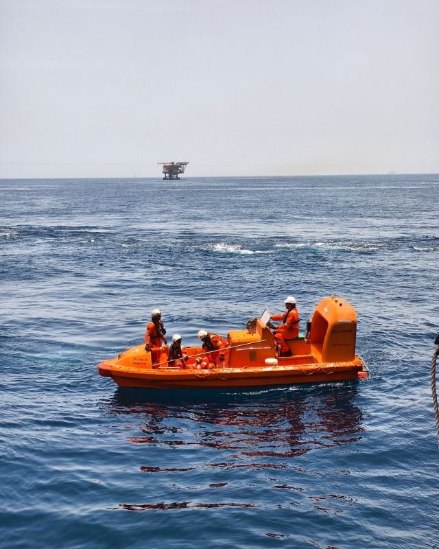 Crew in an orange survival craft during an offshore safety drill, with a platform on the horizon